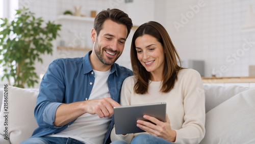 Happy young couple using digital tablet on sofa. Smiling man and woman looking at screen in modern living room. Online shopping and entertainment concept