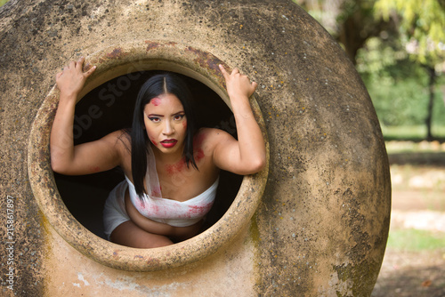 A young Latina woman with dark skin and red eyes, her body wrapped in bandages and covered in wounds, emerges from inside a huge clay jar in a post-apocalyptic city, as if she had turned into a zombie