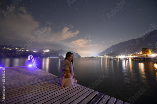 Foto Woman sits on dock at night by lake with stars and lights reflected on water