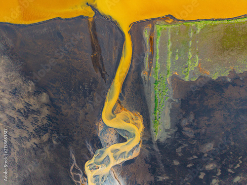 Aerial view of a vibrant yellow river delta winding through dark volcanic terrain and patches of green moss in Iceland.