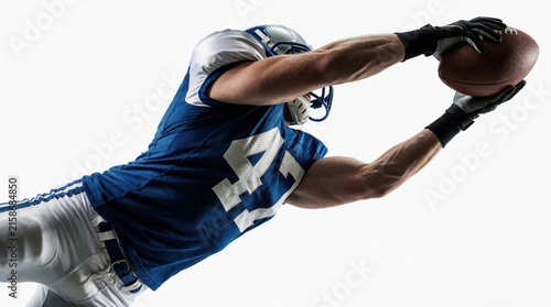 American football player jumps to catch ball in air. Athlete wears blue jersey, helmet during intense match. Pro sportsman reaches for pigskin on white background. Dynamic action shot of male player