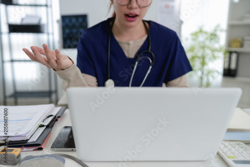 Young Caucasian female doctor in blue scrubs gesturing while conducting online telemedicine consultation on laptop