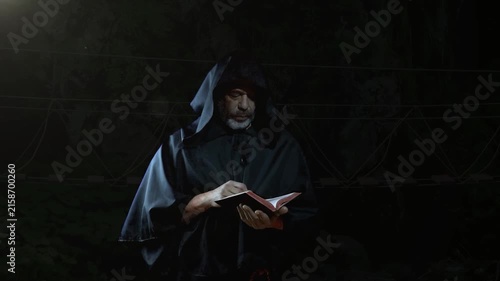 Hooded mature man in dark cloak opens book and reads solemnly