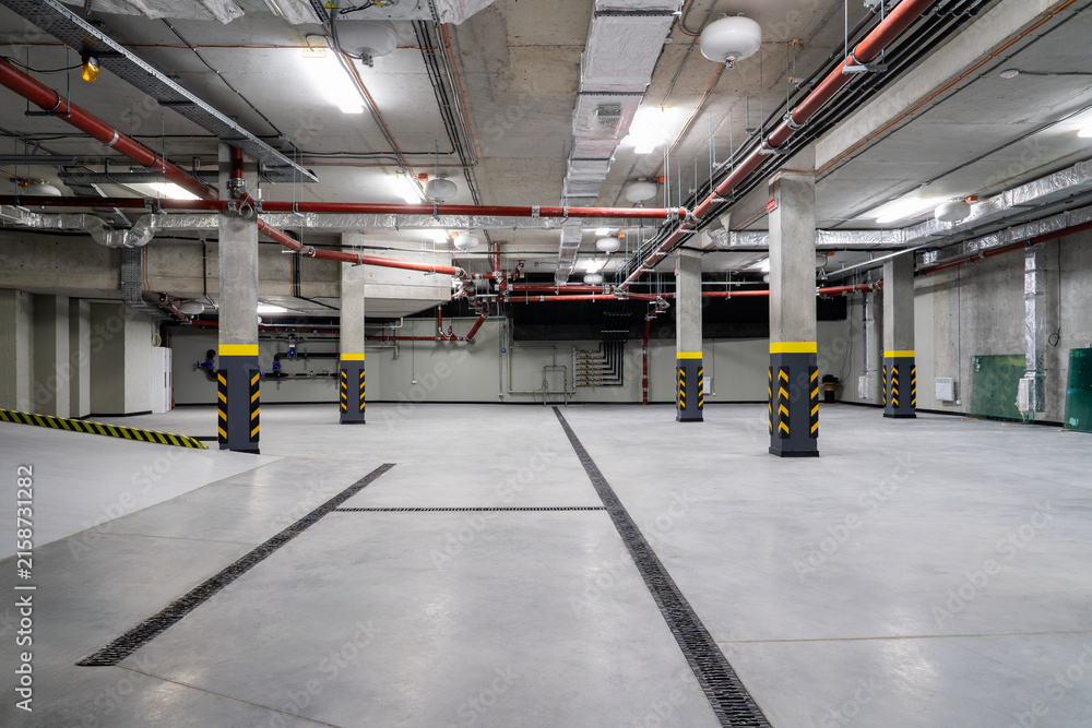 Obraz premium Modern underground parking garage with concrete columns, red fire sprinkler pipes, ventilation ducts and drainage channels in a newly constructed residential building