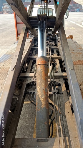 Close-up view of a heavy-duty hydraulic lifting mechanism, showing a large hydraulic cylinder, steel frame, hydraulic lines, and weathered metal components