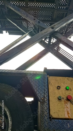 Close-up low-angle view of an old industrial vehicle showing a worn tire, hydraulic lifting mechanism, steel frame, and weathered metal components against the sky