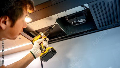 Asian male technician using electric screwdriver fixing extractor motor in cooker hood in modern kitchen. Maintenance worker man repairing exhaust fan in kitchen hood. Home appliance service