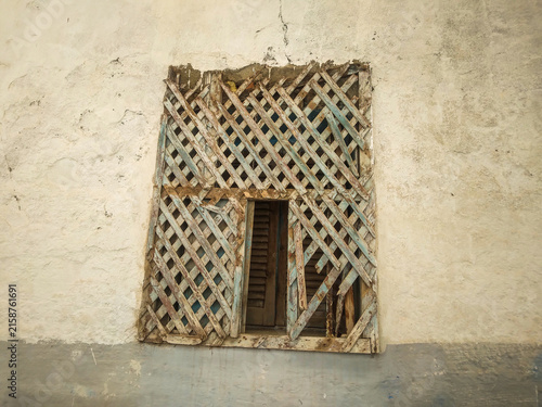 Weathered wooden lattice window screen with peeling blue paint covers a rustic aged stucco wall of an old historic Mediterranean house during a sunny day
