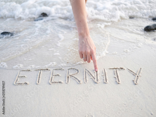 Hand writing the word eternity in wet sand on a beach with ocean waves.