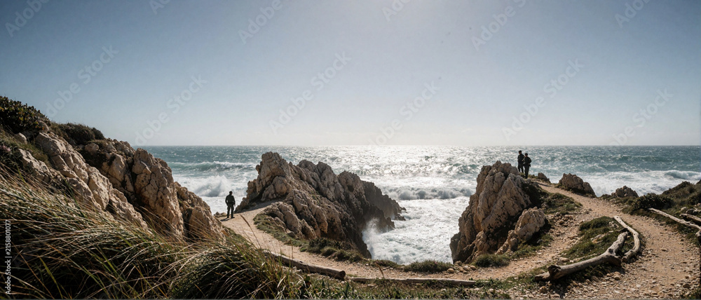 Fototapeta premium Panoramic view of rugged coastal cliffs with people walking on a sandy path overlooking the ocean