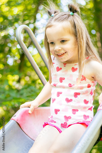 Young girl with long hair wearing a heart-patterned tank top slides down a playground slide surrounded by green trees and sunlight in a park setting