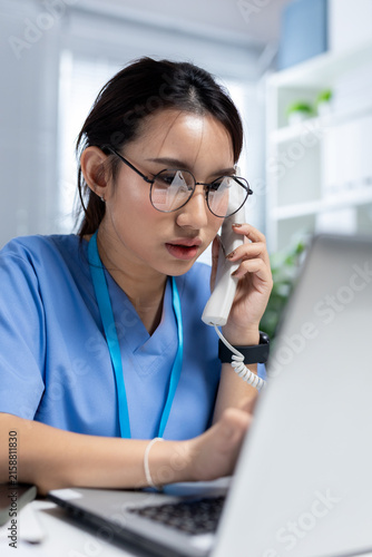 Obraz na plátne women in blue scrubs answering telephone while using laptop in medical office, representing patient support, telehealth service and healthcare communication