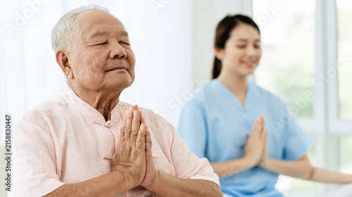 Senior Asian man caregiver practicing meditation with prayerful hands and calm expression in bright care facility