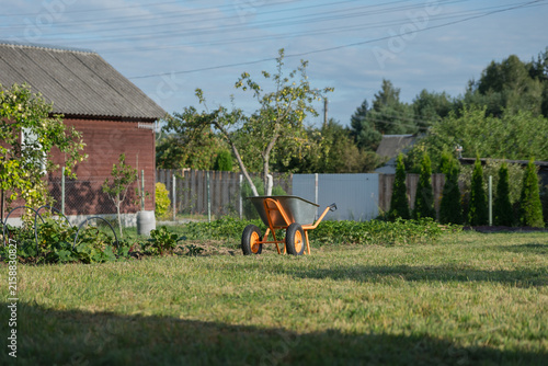 A wooden wheelbarrow stands in a grassy yard beside a rustic house. Trees and shrubs surround the area, creating a peaceful garden scene.