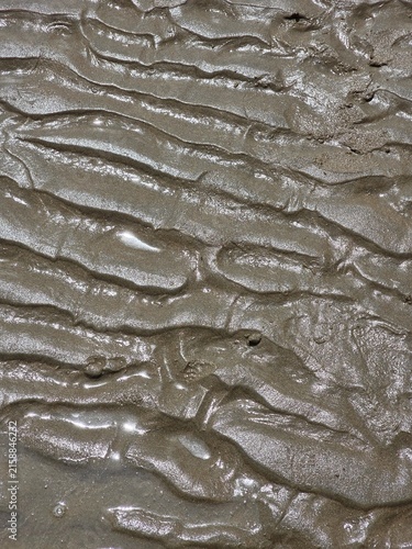 Konstfotografi Textural, high-angle close-up photograph of wet, dark, muddy sand patterns formed by receding tide water on a coastal beach, showing intricate ripples and small puddles