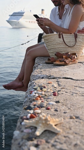 Couple sitting on concrete pier looking at smartphone with ocean cruise ship background vertical