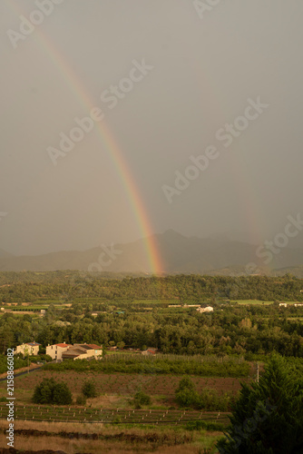 Obraz na plátne A scenic landscape view of a colorful rainbow stretching across a stormy gray sky over green valleys, vineyards, and mountain ranges
