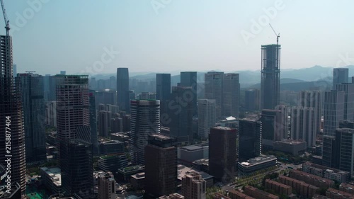 A wide aerial view of a dense modern city skyline with multiple high-rise buildings, some under construction, set against a hazy sky and distant hills.