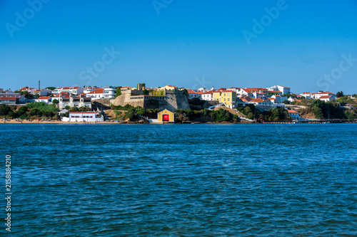 Wide view of the town of Vila Nova de Milfontes from across the Mira River, showing the historic Forte de São Clemente fortress, the colorful lifesaving station at its base, and whitewashed houses 