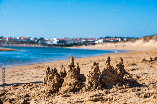 Close-up of textured drip-sand castle towers built on a sandy beach, with calm blue water and a whitewashed coastal town blurred in the background under a clear sky.