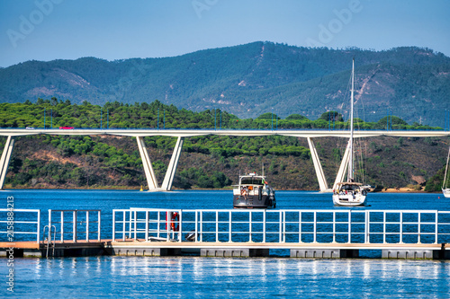 A modern road bridge spans a wide river lined with pine-covered hills, with a motor yacht and sailboat anchored near a floating pier in the foreground, under a clear blue sky in the Alentejo