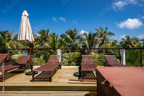 Canvastavla Wooden sun loungers and a parasol stand on an outdoor terrace overlooking tropical palm trees under a bright sunny sky on Dhigurah island, Maldives