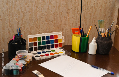 Art supplies are arranged on a wooden desk with paper, paints, and brushes in a creative workspace