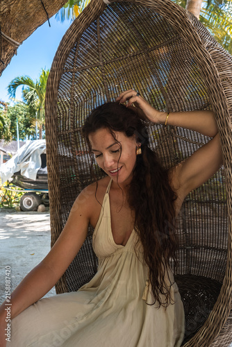Bild auf Leinwand A smiling woman in a beige sundress sits comfortably in an egg shaped woven wicker hanging chair on a tropical island beach in Dhigurah, Maldives