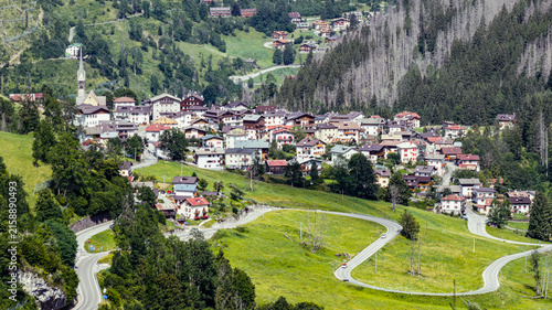 Aerial view of the village of Col di Rocca nestled in the Dolomites with traditional houses, a church spire, winding mountain roads, and lush green meadows in Col di Rocca, Veneto, Italy.