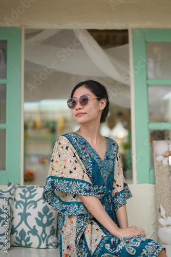 Cuadro en lienzo Asian woman in a batik kaftan and sunglasses relaxing at a beach cafe