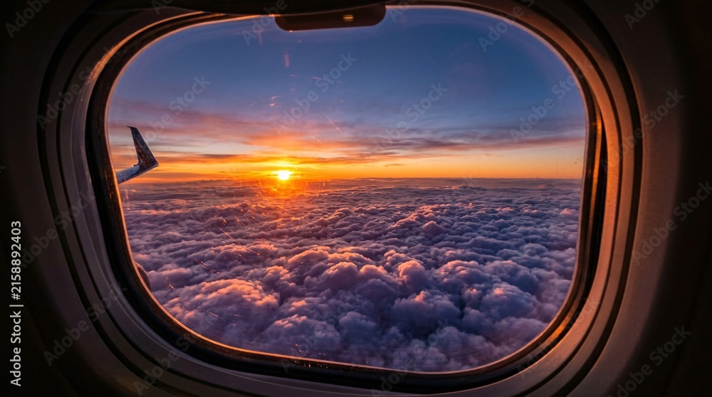 Fototapeta premium Airplane window frames a mesmerizing high-altitude sunset looking down upon a dense fluffy sea of white and purple clouds glowing brightly.
