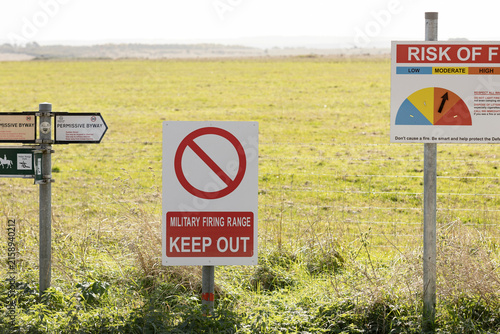 A military firing range 'Keep Out' sign with a nearby risk of fire notice mounted on a fence beside open grassland, warning the public of restricted access and potential hazards in the countryside.