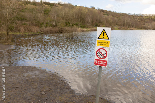 A warning sign reading 'Dangerous Water' and 'Do Not Enter The Water' stands at the edge of a flooded lake shore, conveying caution, restricted access and public safety in a rural landscape.