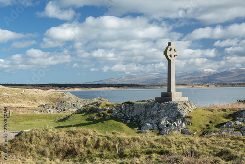 A solitary Celtic cross memorial stands on rocky grassland above a calm sea, framed by rolling dunes, distant mountains and a bright sky with scattered clouds, 