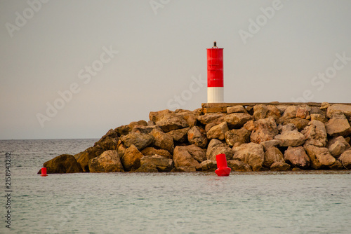 Red and white harbor beacon light standing on a rocky breakwater pier surrounded by calm sea water at dusk.

