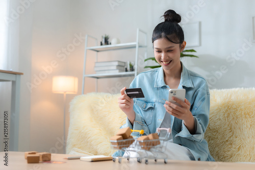 Happy woman shopping online with smartphone and credit card at home, using e-commerce and mobile payment with miniature carts, packages for convenient digital retail delivery.