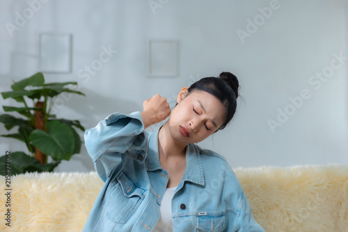 Young woman suffering from neck pain while sitting on sofa at home, representing muscle tension, stiffness, fatigue, discomfort, poor posture, stress and everyday wellness concerns.