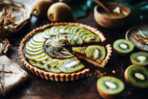 A kiwi and mint pie is placed on a brown plate. It is topped with thin slices of kiwi, cookie pieces, micro herbs, and a green foam sauce. The scene shows a cafe setting with soft lighting
