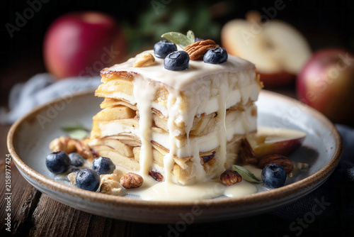 Layered apple pie sits on a wooden table. The pie shows layers of dough and fruit. Soft shadows and highlights appear from natural light