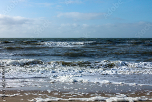 Waves leave behind white foam in the foreground, spreading onto the brown sand of the beach, under a blue sky dotted with a few scattered clouds on a sunny day