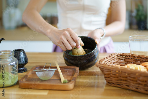 Asian woman prepares matcha tea by whisking green powder in a bowl with a bamboo whisk, showcasing the blending process in a bright kitchen setting
