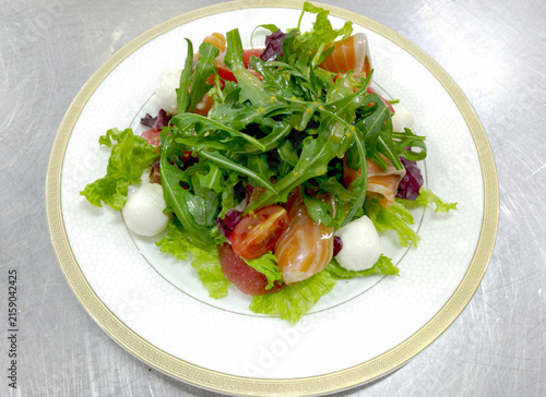 A fresh salad featuring arugula, tomato pieces, slices of lightly salted red fish, and mozzarella balls. It is complemented by juicy vegetables and a light dressing. The dish looks appetizing.
