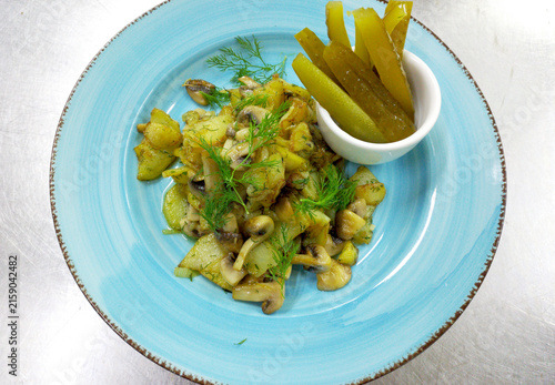 Fried potatoes with mushrooms and onions. Served with pickles on a blue plate. Garnished with sprigs of fresh dill. Delicious food, Russian culture.
