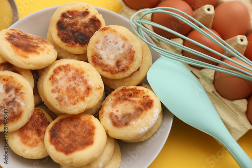 Homemade delicious syrniki, cheese pancakes in plate, cooking ingredients, kitchen utensils on yellow background