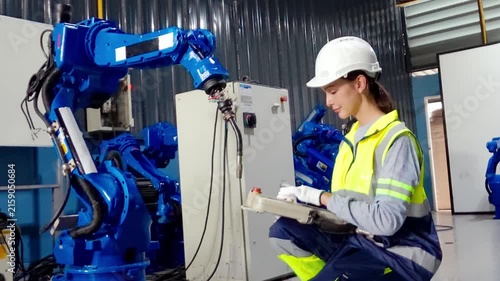 Robotic Arm Operation: A female engineer in a hard hat and safety vest works with a sophisticated blue robotic arm on an industrial floor, controlling it with a console.