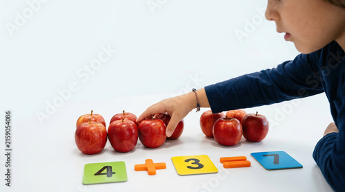 Child demonstrating addition with apples and number tiles at white table, focused on early mathematics learning through colorful hands on activity