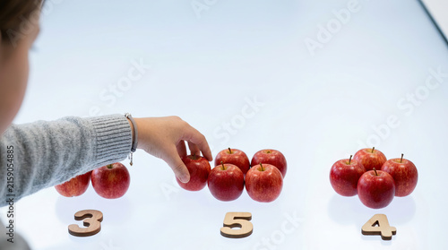 Student counting apple group on white table with wooden number marker, learning arithmetic through visual comparison and focused classroom activity