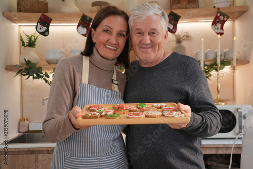 An elderly couple, smiling happily, dressed in aprons, carried wooden trays and baked and decorated gingerbread cookies together to celebrate Christmas with their family.