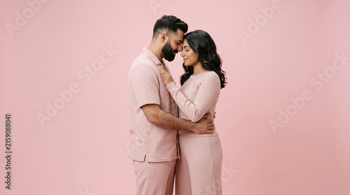 Romantic couple in matching pink outfits embracing and smiling at each other against a soft pastel pink studio background.