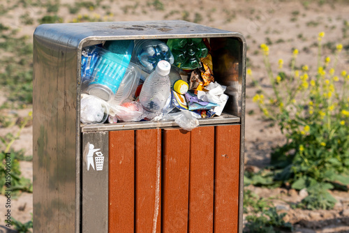 A modern outdoor public trash can overflowing with discarded plastic bottles, food wrappers, and single-use packaging. 
